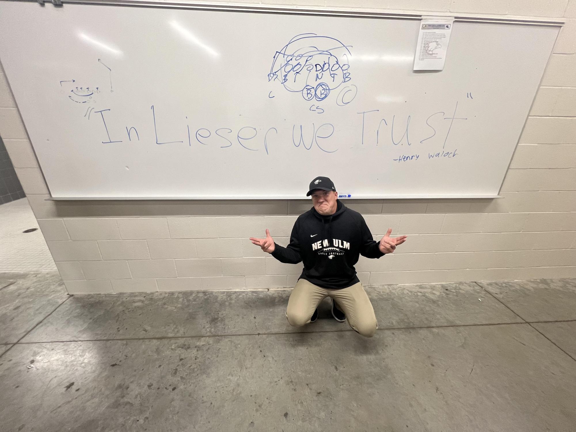 Football coach Derek Lieser poses in front of the white board with a hopeful slogan on it: In Lieser we Trust.